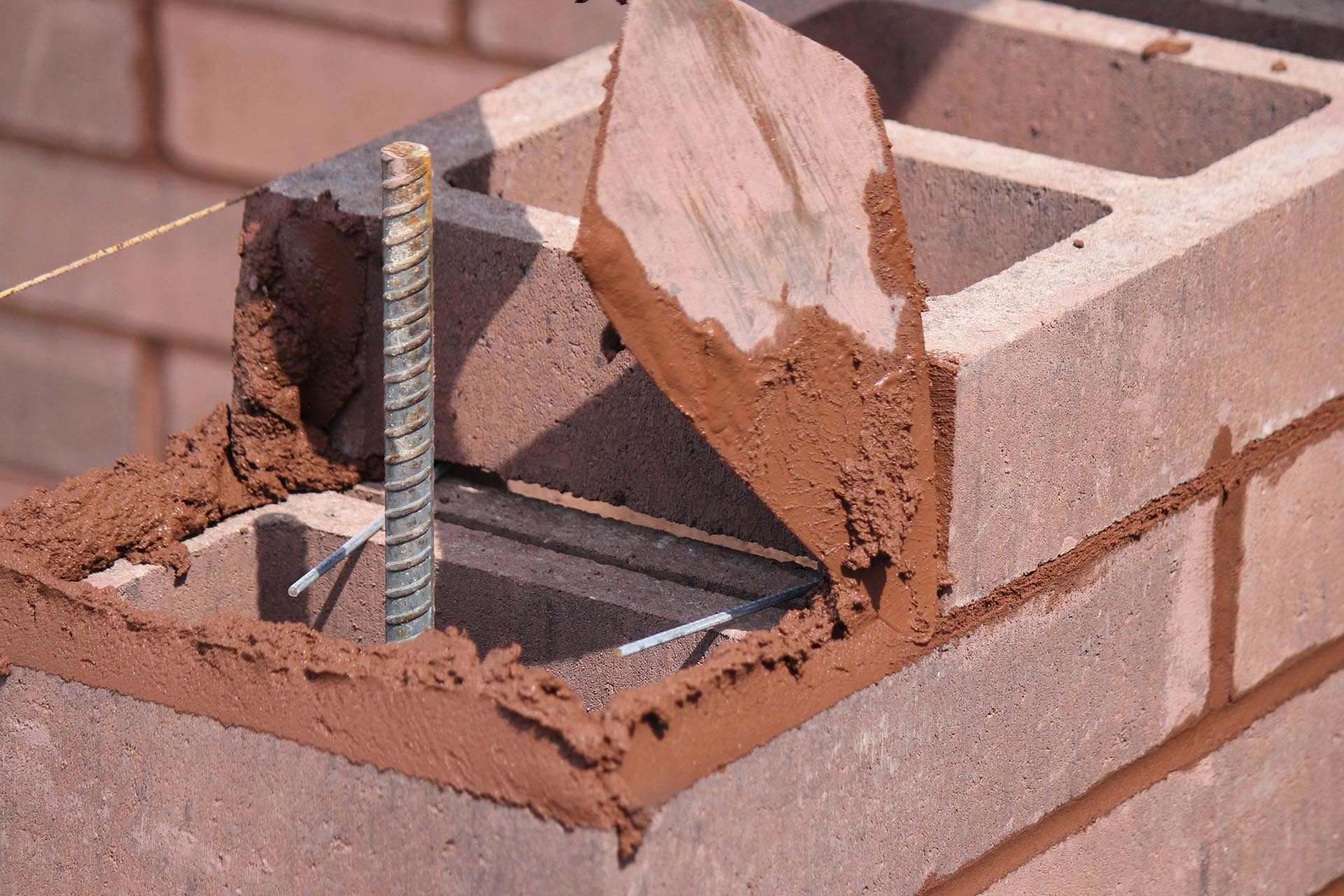 A mason using a trowel to set a colored concrete block in a wall being built, with an exposed rebar protruding above the block wall.